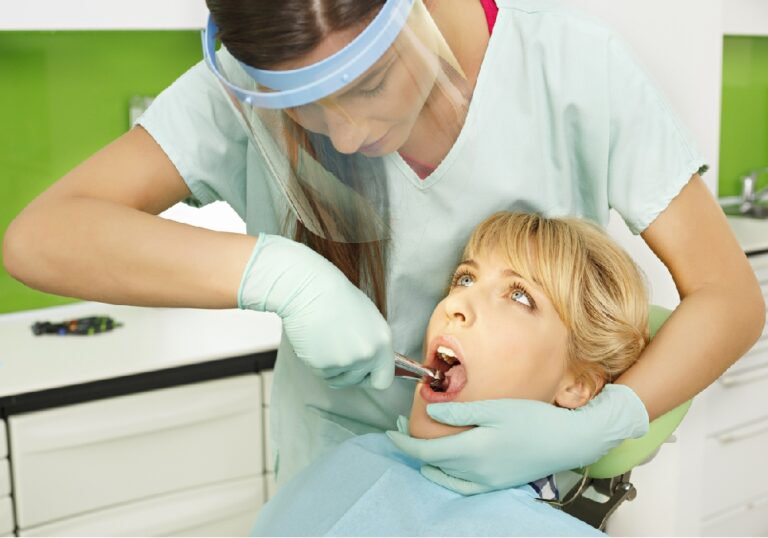 A dental professional wearing gloves and a protective face shield is performing a dental procedure on a patient seated in a dental chair. Dental equipment and cabinetry are visible in the background.