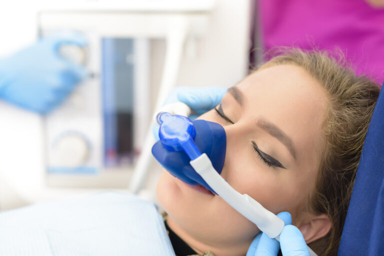 A woman in a medical setting is inhaling sedation through a mask, while a gloved hand adjusts equipment in the background.